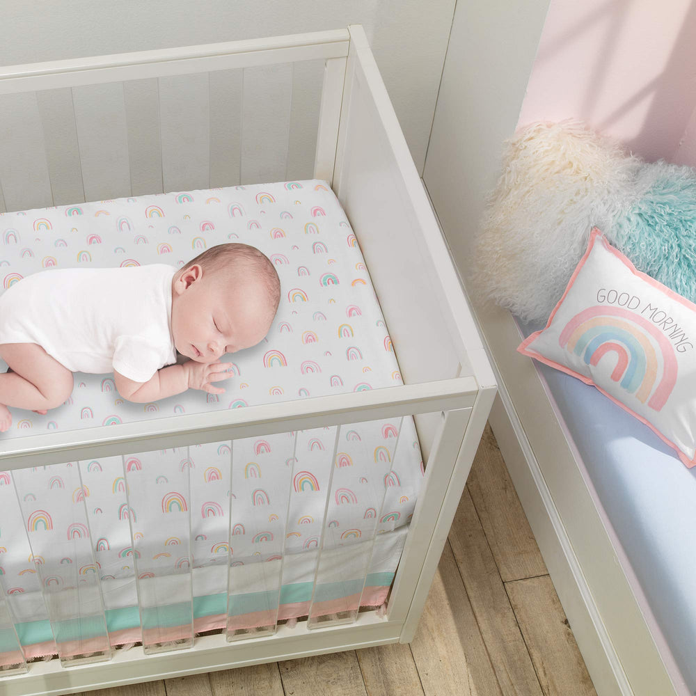 Lambs & Ivy Watercolor Rainbow sheet flat lay highlights pastel rainbows on white ground for nursery decor.